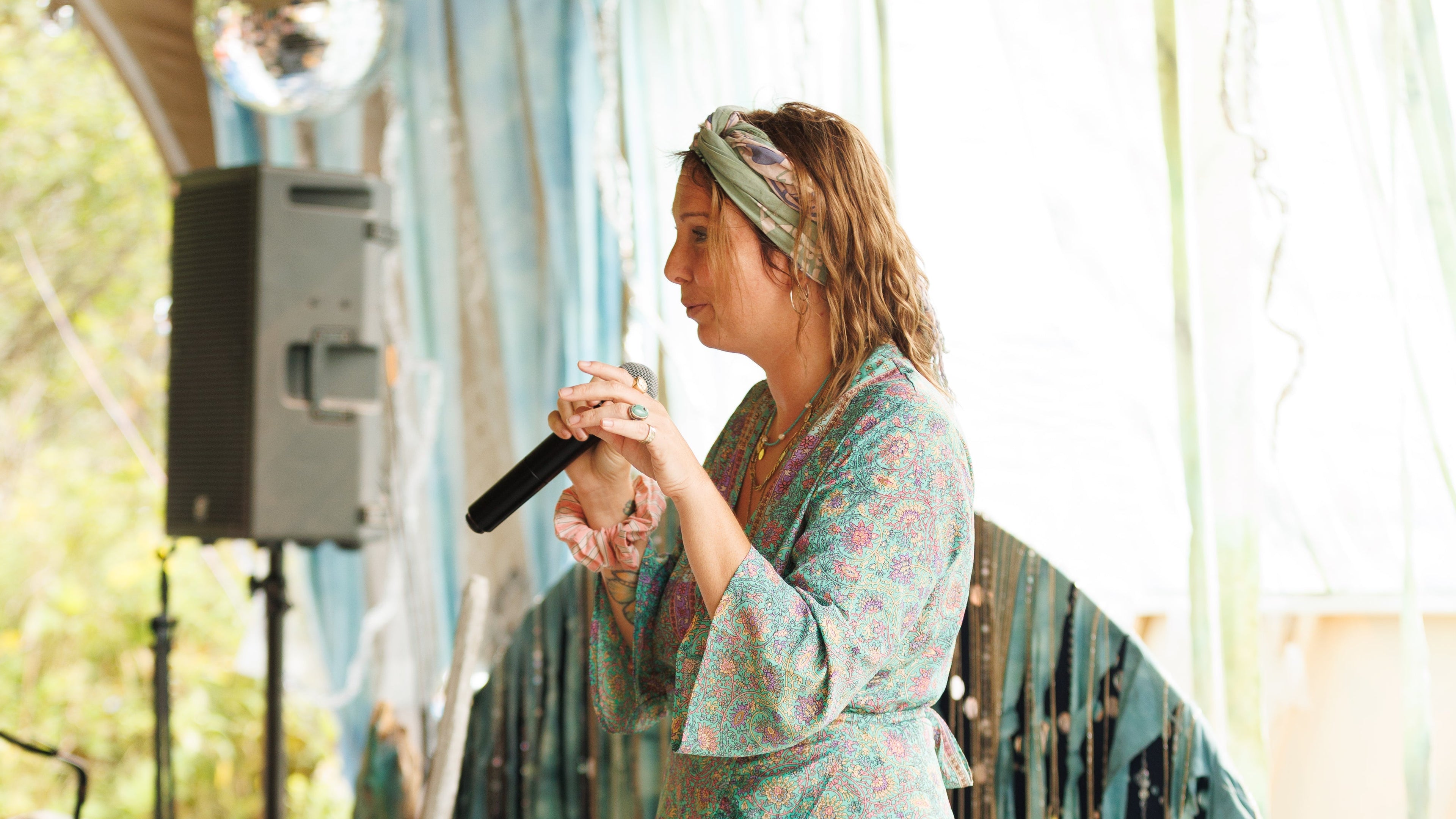 Conference Anatha. Woman in a floral dress standing in a tent-like structure with curtains and a microphone.